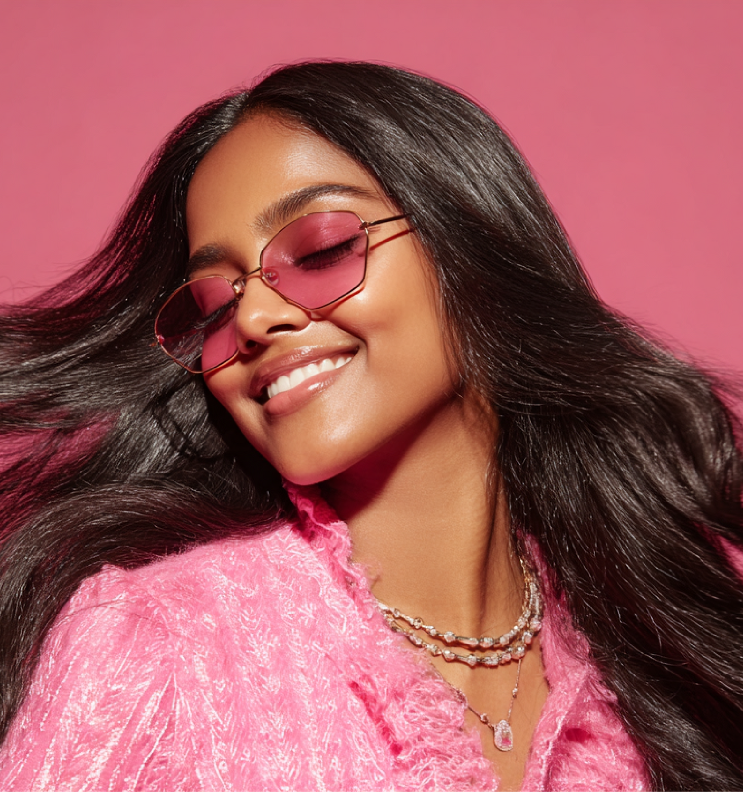 Woman with long dark hair wearing pink-tinted sunglasses and a textured pink top smiling with eyes closed against a pink background
